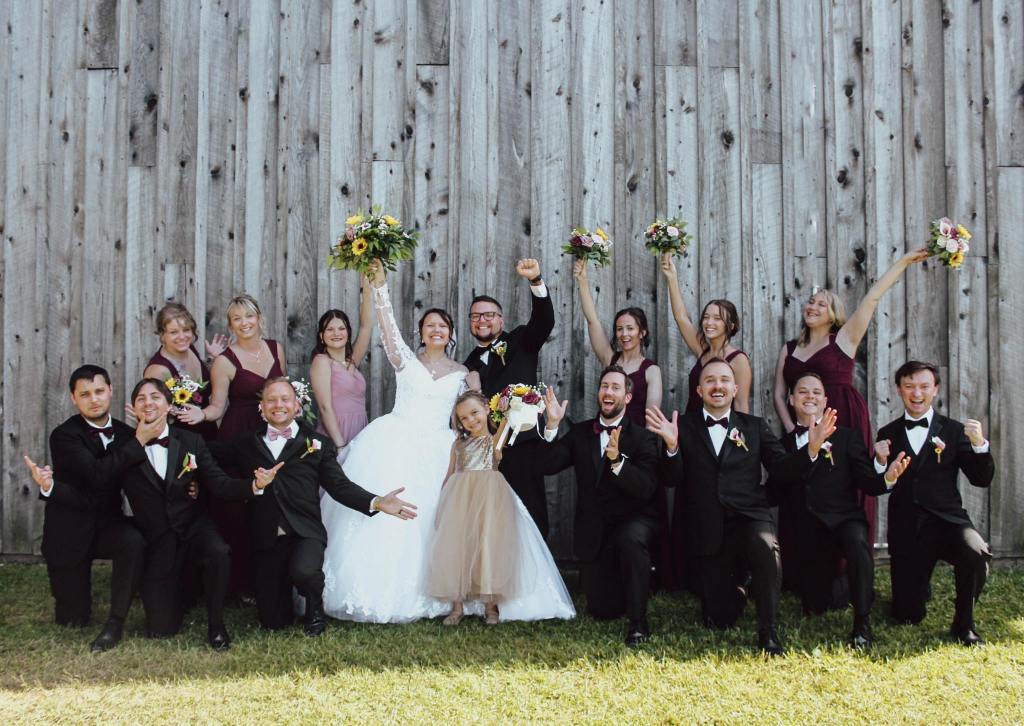 The image depicts a joyous wedding party, with the members of the bridal party appearing visibly elated and enthusiastic. The individuals in the photograph are clearly celebrating the momentous occasion, their expressions radiating a sense of excitement and happiness. The overall composition and body language of the group conveys the celebratory atmosphere and the shared delight experienced by those in attendance.
